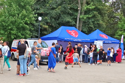 Na tle głównym stoisko SPZZLO Warszawa Żoliborz-Bielany z pacjentami ustawionymi w kolejce.