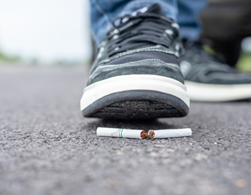 Na zdjęciu widać zbliżenie buta sportowego, który przygniata niedopałek papierosa leżący na asfaltowej nawierzchni.